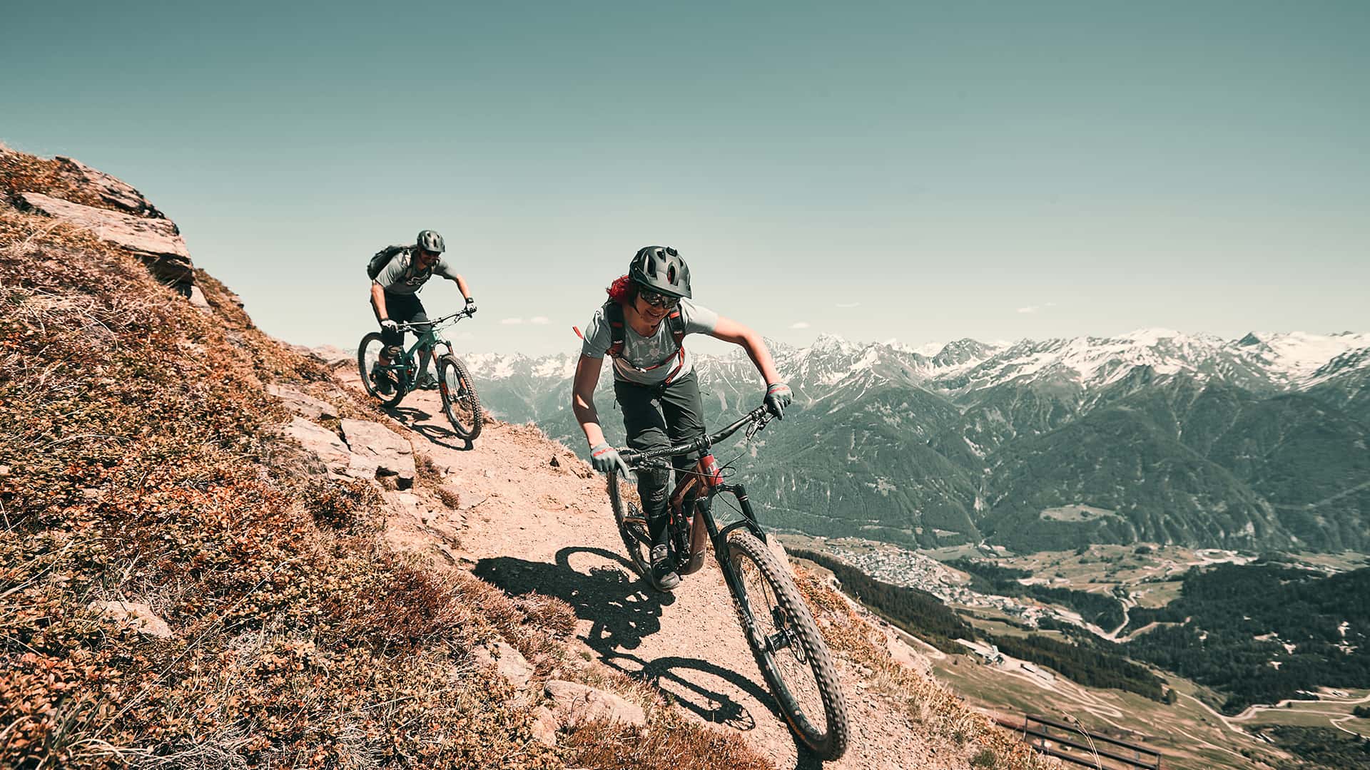 Zwei Personen fahren auf ihrem Mountainbike einen Hang hinunter. Im Hintergrund ist ein Panorama über die Tiroler Alpen zu sehen.